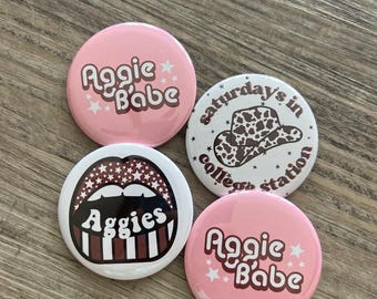 Op de afbeelding: Vier ronde roze en witte knoppen met verschillende ontwerpen. Eén knop zegt "Aggie Babe" in een retro-lettertype met witte sterren. Een andere knop zegt "Aggie Babe" in een retro-lettertype met witte sterren. Een andere knop zegt "Saturdays in College Station" met een hoed met een koe-print. De laatste knop heeft rode en witte gestreepte lippen met het woord "Aggies" in een retro-lettertype.
