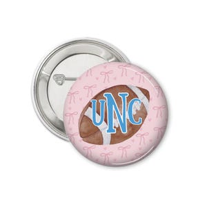 Peut inclure: Un bouton rond avec un fond rose repr&eacute;sentant un ballon de football aquarelle. Le ballon est marron avec des rayures blanches et les lettres "UNC" en bleu. Le fond pr&eacute;sente un motif de petits n&oelig;uds roses.