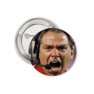 May include: A round button featuring a close-up of a man with an open mouth, wearing a headset with a microphone. The button has a silver metal backing.