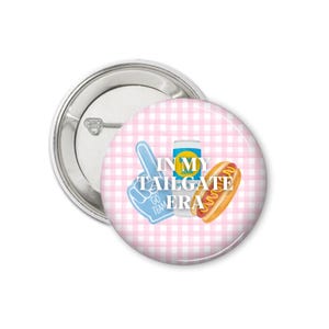 Peut inclure: Un bouton rond avec une &eacute;pingle argent&eacute;e. Le bouton pr&eacute;sente un motif vichy rose et blanc. Des graphiques incluent un doigt en mousse bleu, une canette de soda et un hot-dog. Le texte dit "IN MY TAILGATE ERA."