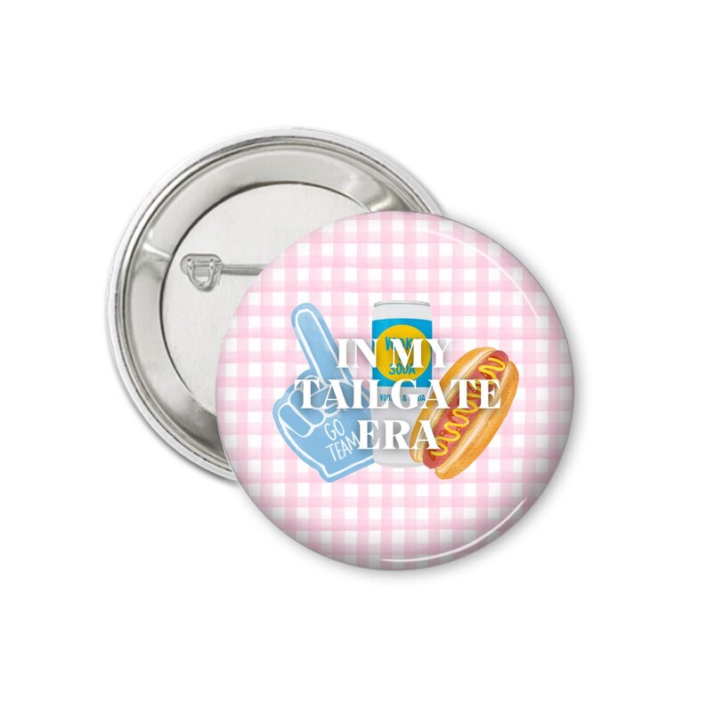Peut inclure: Un bouton rond avec une &eacute;pingle argent&eacute;e. Le bouton pr&eacute;sente un motif vichy rose et blanc. Des graphiques incluent un doigt en mousse bleu, une canette de soda et un hot-dog. Le texte dit "IN MY TAILGATE ERA."