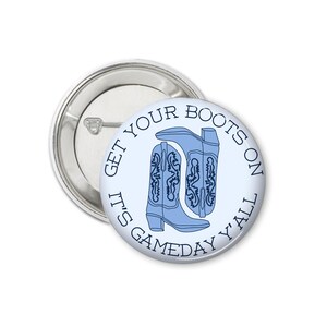 Peut inclure: Un bouton bleu clair avec le texte "GET YOUR BOOTS ON IT'S GAMEDAY Y'ALL" encerclant une paire de bottes de cowboy bleu clair. Le bouton est rond et poss&egrave;de une &eacute;pingle argent&eacute;e.
