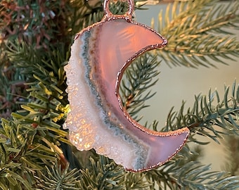 May include: A pink and green agate crescent moon ornament with a copper wire frame. The ornament is hanging from a brown twine string on a green artificial Christmas tree.