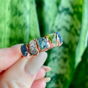 May include: A rose gold ring with five raw gemstones set in a band. The stones are green, blue, and gray.