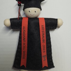 Pode incluir: Um ornamento de formatura com uma figura de madeira vestindo um capelo e uma beca de feltro preto. Duas faixas vermelhas pendem na frente, impressas com o texto "TEXAS TECH UNIVERSITY" e "LEO STRAUBE - 2023" junto com um logotipo universit&aacute;rio.
