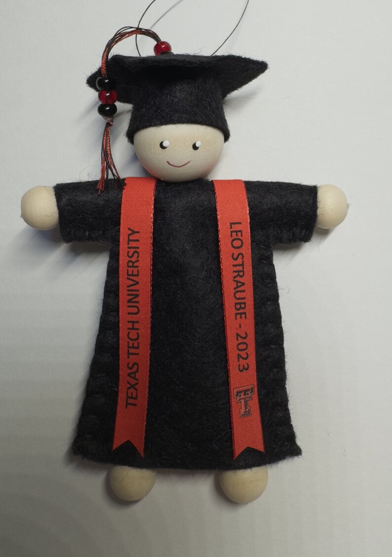 Pode incluir: Um ornamento de formatura com uma figura de madeira vestindo um capelo e uma beca de feltro preto. Duas faixas vermelhas pendem na frente, impressas com o texto "TEXAS TECH UNIVERSITY" e "LEO STRAUBE - 2023" junto com um logotipo universit&aacute;rio.