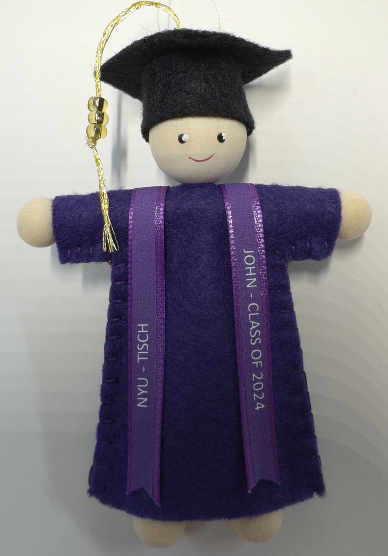 Pode incluir: Um ornamento de formatura com uma boneca de madeira usando um capelo de feltro preto com borla dourada e uma beca de feltro roxa. Duas fitas roxas pendem na frente, impressas com o texto NYU - TISCH e JOHN - CLASS OF 2024.