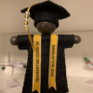 Pode incluir: Um ornamento suspenso em forma de graduado usando um capelo e uma beca de feltro preto. Duas fitas douradas pendem na frente, com o texto QUARANTINE COVID-19 &agrave; esquerda e GRADUATION 2020 &agrave; direita. Um pend&atilde;o dourado com contas est&aacute; anexado.