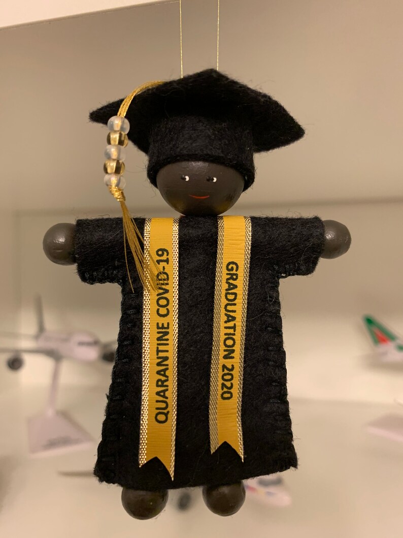 Pode incluir: Um ornamento suspenso em forma de graduado usando um capelo e uma beca de feltro preto. Duas fitas douradas pendem na frente, com o texto QUARANTINE COVID-19 &agrave; esquerda e GRADUATION 2020 &agrave; direita. Um pend&atilde;o dourado com contas est&aacute; anexado.