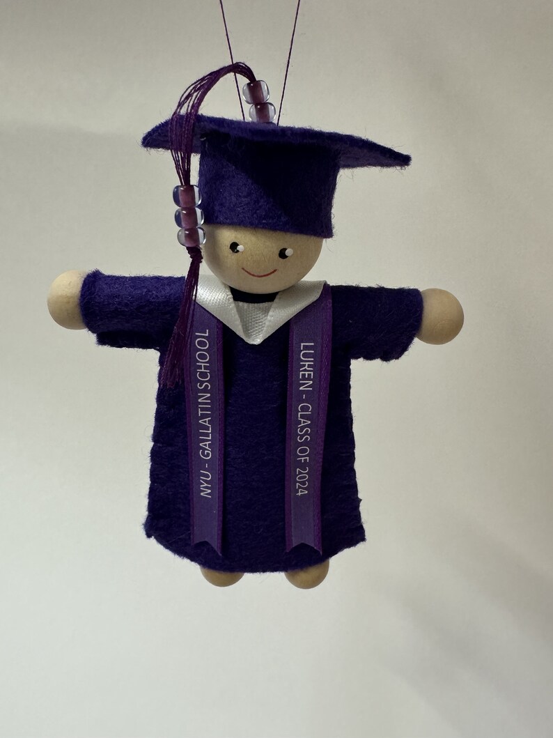 Pode incluir: Um ornamento de formatura com uma boneca de madeira vestindo um capelo e beca de feltro roxo. Uma borla roxa com tr&ecirc;s contas transparentes pende do capelo. Um colarinho branco e uma estola roxa trazem impresso NYU - GALLATIN SCHOOL e LUKEN - CLASS OF 2024.