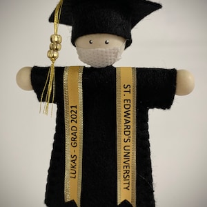 Pode incluir: Um ornamento de formatura com uma figura de madeira vestindo um capelo e beca de feltro preto, uma m&aacute;scara facial branca e duas faixas douradas. Uma faixa diz LUKAS - GRAD 2021 e a outra diz ST. EDWARD'S UNIVERSITY. Um pend&atilde;o dourado pende.