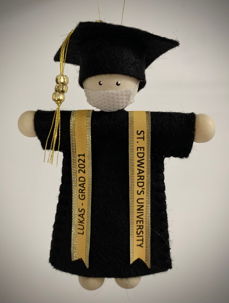 Pode incluir: Um ornamento de formatura com uma figura de madeira vestindo um capelo e beca de feltro preto, uma m&aacute;scara facial branca e duas faixas douradas. Uma faixa diz LUKAS - GRAD 2021 e a outra diz ST. EDWARD'S UNIVERSITY. Um pend&atilde;o dourado pende.