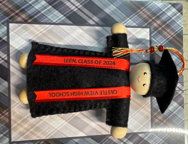Pode incluir: Um ornamento de formatura com uma cabe&ccedil;a de conta de madeira com rosto pintado, usando um capelo e uma beca de feltro preto. Duas fitas vermelhas est&atilde;o presas &agrave; beca, impressas com o texto LEEN, CLASS OF 2024 e CASTLE VIEW HIGH SCHOOL.
