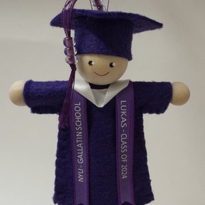 Pode incluir: Um ornamento de formatura com uma boneca de madeira vestindo um capelo e uma beca de feltro roxo. A boneca tem uma borla roxa com contas e um colarinho branco. Duas fitas roxas pendem com o texto NYU - GALLATIN SCHOOL e LUKAS - CLASS OF 2024.