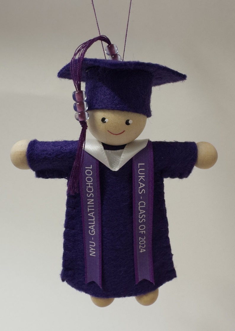 Pode incluir: Um ornamento de formatura com uma boneca de madeira vestindo um capelo e uma beca de feltro roxo. A boneca tem uma borla roxa com contas e um colarinho branco. Duas fitas roxas pendem com o texto NYU - GALLATIN SCHOOL e LUKAS - CLASS OF 2024.