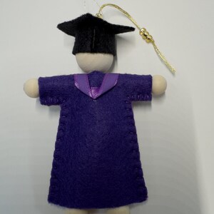 Pode incluir: Um ornamento de formatura com uma figura de madeira vestindo uma toga de feltro roxa com costuras vis&iacute;veis e um colarinho de fita roxa. A figura usa um capelo de feltro preto e tem um cord&atilde;o dourado com tr&ecirc;s pequenas contas douradas.