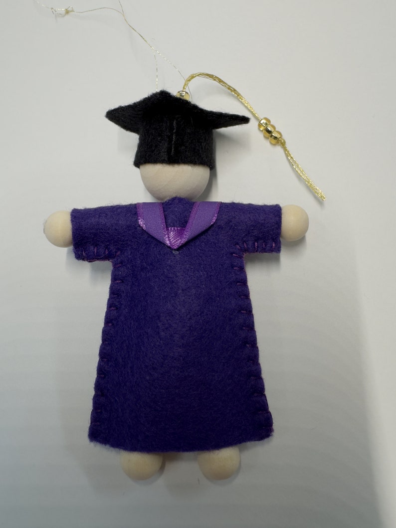 Pode incluir: Um ornamento de formatura com uma figura de madeira vestindo uma toga de feltro roxa com costuras vis&iacute;veis e um colarinho de fita roxa. A figura usa um capelo de feltro preto e tem um cord&atilde;o dourado com tr&ecirc;s pequenas contas douradas.