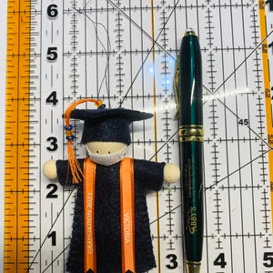 Pode incluir: Um ornamento de formatura em feltro com uma figura de capelo e toga azul-escura e uma m&aacute;scara facial branca. Duas fitas laranja est&atilde;o presas, uma com "GRADUATION 2021" e a outra com "VICTORIA". Uma r&eacute;gua mostra 8,9 cm de altura.
