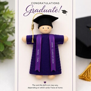 Pode incluir: Uma boneca de madeira com um capelo de gradua&ccedil;&atilde;o de feltro preto com borla dourada e uma toga de feltro roxa. Duas fitas roxas pendem dos ombros, impressas com "NYU - TISCH" e "JOHN - CLASS OF 2024". A boneca est&aacute; montada num cart&atilde;o branco.