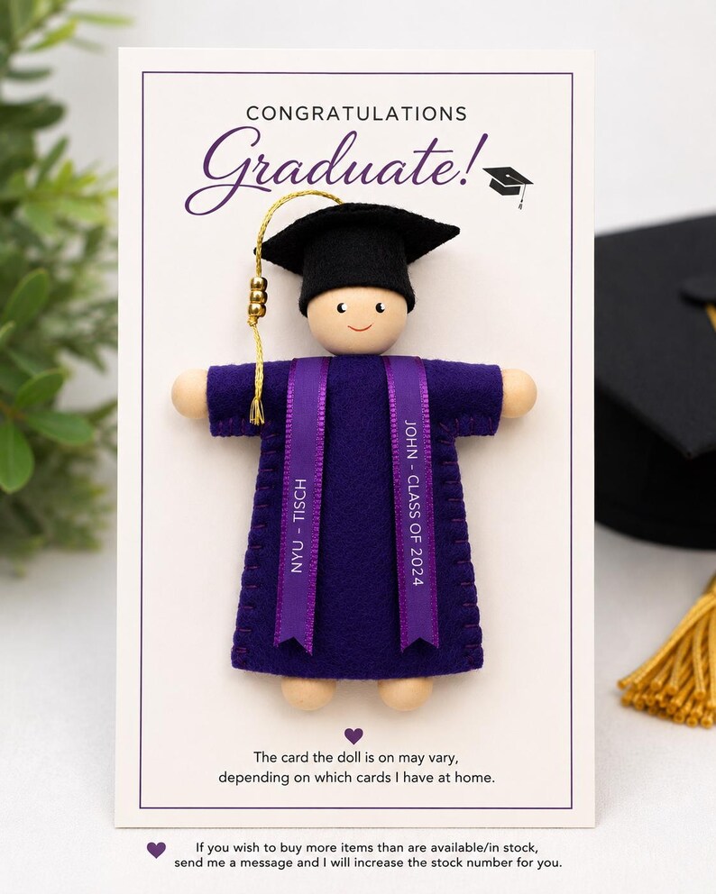 Pode incluir: Uma boneca de madeira com um capelo de gradua&ccedil;&atilde;o de feltro preto com borla dourada e uma toga de feltro roxa. Duas fitas roxas pendem dos ombros, impressas com "NYU - TISCH" e "JOHN - CLASS OF 2024". A boneca est&aacute; montada num cart&atilde;o branco.