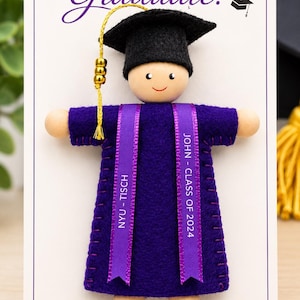 Pode incluir: Um ornamento de formatura com uma boneca de madeira usando um capelo de feltro preto e uma beca de feltro roxa. A beca tem duas fitas roxas com o texto JOHN - CLASS OF 2024 e NYU - TISCH. Uma borla dourada pende do capelo. O cart&atilde;o diz CONGRATULATIONS Graduate!