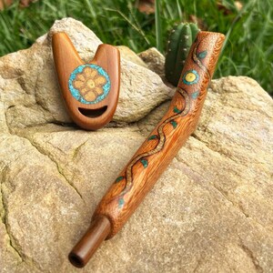 May include: Two wooden pipes rest on a rock. One is a small, light brown pipe with a turquoise and orange floral design. The other is a longer, curved pipe with a brown vine and leaf design. The pipes are handcrafted.