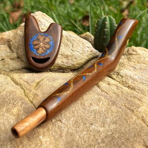 Può includere: Due pipe in legno fatte a mano. Una è una pipa curva con intarsi blu e oro. L'altra è una piccola pipa a forma di U con un motivo floreale e intarsi blu. Entrambe le pipe sono su una grande roccia chiara.