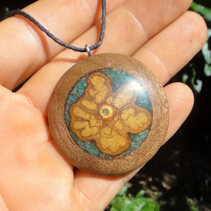 May include: A round wooden pendant with a yellow flower design embedded in resin. The flower has a green and brown marbled pattern. The pendant is on a black cord.