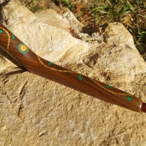 May include: A wooden pipe with a curved design, decorated with inlaid turquoise and gold accents. The pipe is resting on a rock.