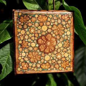 May include: Square wooden art piece with a floral design. The artwork features a mosaic of small, circular wood pieces in shades of brown and yellow, set against a teal background. The central flower is larger and more detailed.