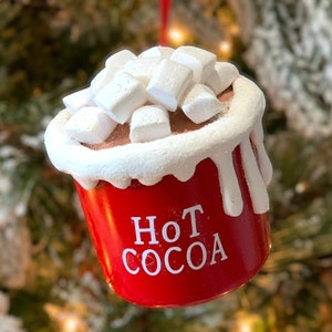 May include: A red ornament shaped like a cup of hot cocoa, topped with white marshmallows. The words "Hot Cocoa" are printed in white on the front of the cup. The ornament has a white rim and white drips down the side.