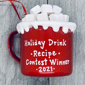 May include: A red ceramic mug ornament with white text that reads "Holiday Drink Recipe Contest Winner 2021." The mug is topped with white, snowy frosting and white marshmallows. A red ribbon is attached for hanging.