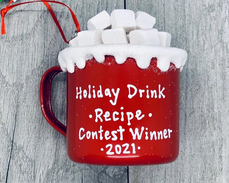 May include: A red ceramic mug ornament with white text that reads "Holiday Drink Recipe Contest Winner 2021." The mug is topped with white, snowy frosting and white marshmallows. A red ribbon is attached for hanging.