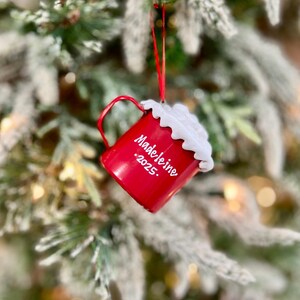 May include: A red Christmas ornament shaped like a mug, hanging from a red ribbon on a Christmas tree. The mug has a white foam-like topping and the name "Madeleine .2025." written on the side. The tree has frosted green needles.