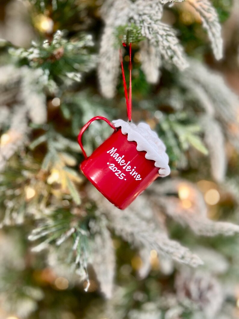 May include: A red Christmas ornament shaped like a mug, hanging from a red ribbon on a Christmas tree. The mug has a white foam-like topping and the name "Madeleine .2025." written on the side. The tree has frosted green needles.