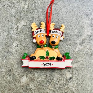 May include: A Christmas ornament featuring two reindeer with the names "Jordan" and "Isabella" on their hats. The reindeer are brown with red noses and green collars. A banner at the bottom reads "2024". The ornament has a red ribbon for hanging.
