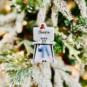 May include: A white Christmas ornament with the name "Jessie" and the year "2024" on it. The ornament depicts a person sitting at a desk. The ornament is hanging on a snow-covered Christmas tree.