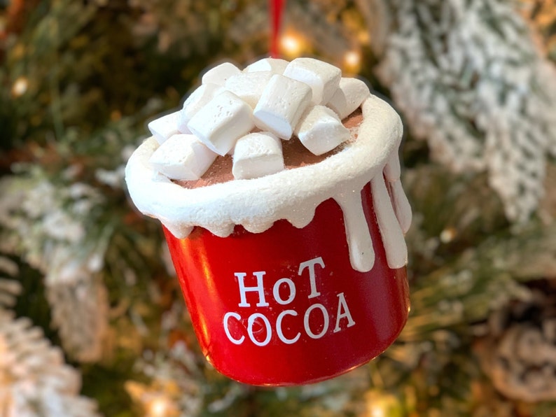May include: A red ornament shaped like a cup of hot cocoa, topped with white marshmallows. The words "Hot Cocoa" are printed in white on the front of the cup. The ornament has a white rim and white drips down the side.