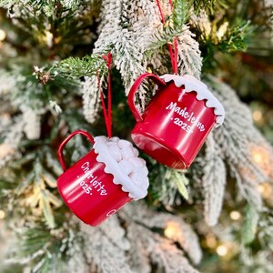 May include: Two red Christmas ornaments shaped like mugs, hanging on a decorated tree. Each mug has a white, fluffy rim and a handle. One ornament reads "Madeleine 2025" and the other reads "Charlotte 025."