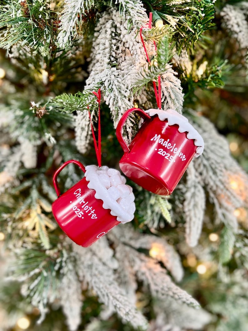 May include: Two red Christmas ornaments shaped like mugs, hanging on a decorated tree. Each mug has a white, fluffy rim and a handle. One ornament reads "Madeleine 2025" and the other reads "Charlotte 025."