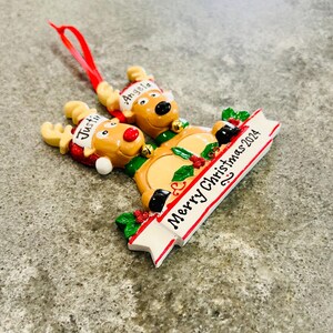 May include: A Christmas ornament featuring three reindeer with the names "Justin" and "Angela" on their hats. The ornament has a banner that reads "Merry Christmas 2024" and is decorated with holly and berries.