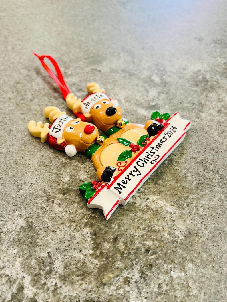 May include: A Christmas ornament featuring three reindeer with the names "Justin" and "Angela" on their hats. The ornament has a banner that reads "Merry Christmas 2024" and is decorated with holly and berries.