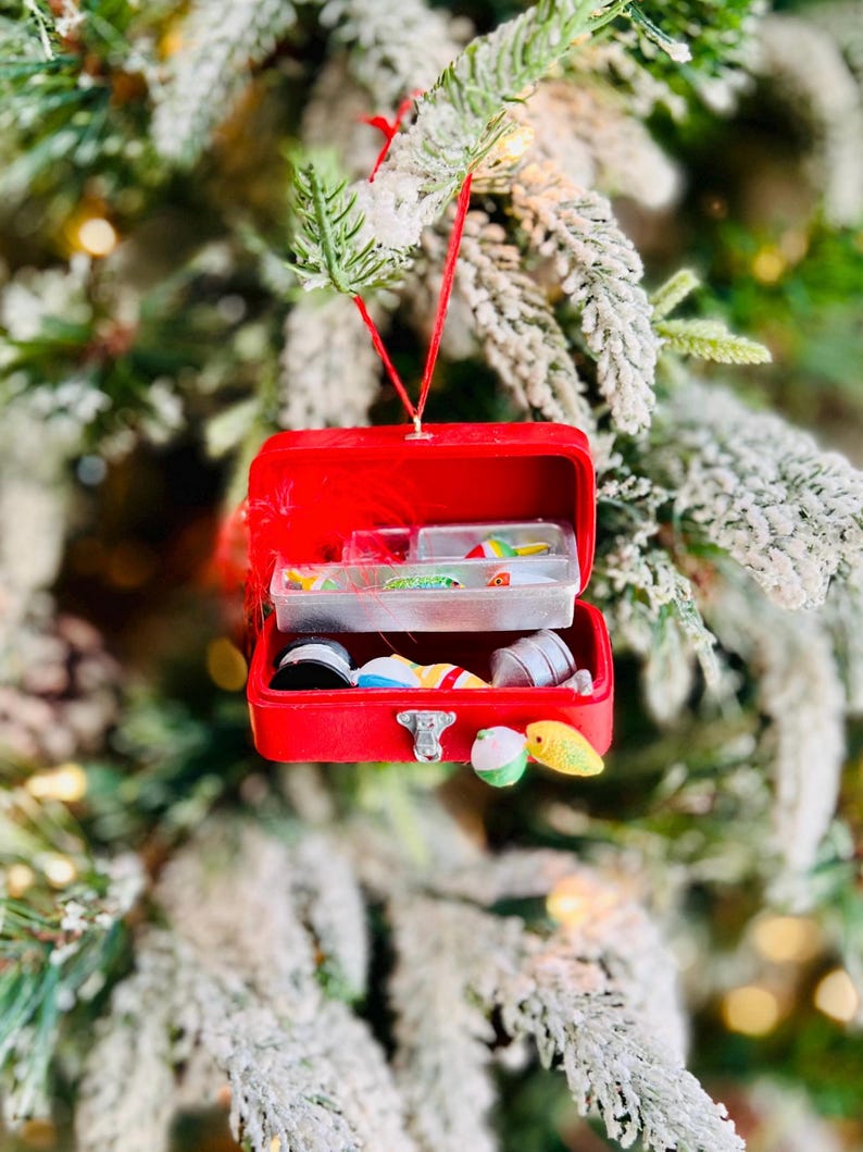 May include: A red Christmas ornament shaped like a miniature tackle box, open to reveal small fishing lures and accessories. The ornament hangs from a red string against a backdrop of a snow-covered Christmas tree.