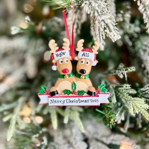 May include: A Christmas ornament featuring two reindeer with the names "Kyle" and "Ali" on their hats. The reindeer have brown bodies, red glitter antlers, and green collars. A banner below reads "Merry Christmas 2025".