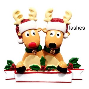 May include: A Christmas ornament featuring two reindeer with tan bodies, wearing Santa hats with red glitter trim and green scarves. The reindeer are positioned above a white banner with a red border, surrounded by holly and berries.