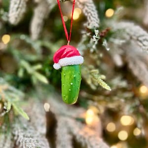 May include: A green pickle Christmas ornament with a red Santa hat and white trim, hanging from a red string. The ornament is detailed with bumps and is set against a blurred Christmas tree background.