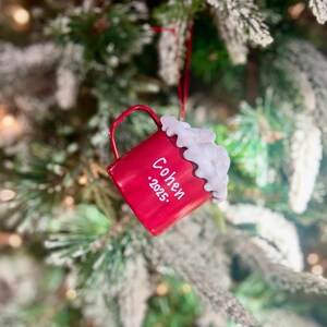 May include: A red Christmas ornament shaped like a mug, with the name "Cohen" and the year ".2025." printed on it. The mug is topped with white, fluffy, whipped cream. The ornament hangs from a red string against a blurred Christmas tree background.