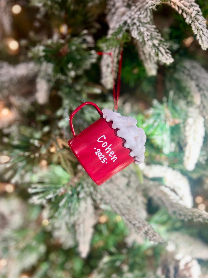 May include: A red Christmas ornament shaped like a mug, with the name "Cohen" and the year ".2025." printed on it. The mug is topped with white, fluffy, whipped cream. The ornament hangs from a red string against a blurred Christmas tree background.