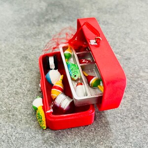 May include: A red miniature tackle box ornament, open to reveal colorful fishing bobbers and lures. The box is rectangular with a hinged lid and a small red ribbon for hanging. The lures include a green and blue fish, a white and red bobber, and a yellow and green lure.