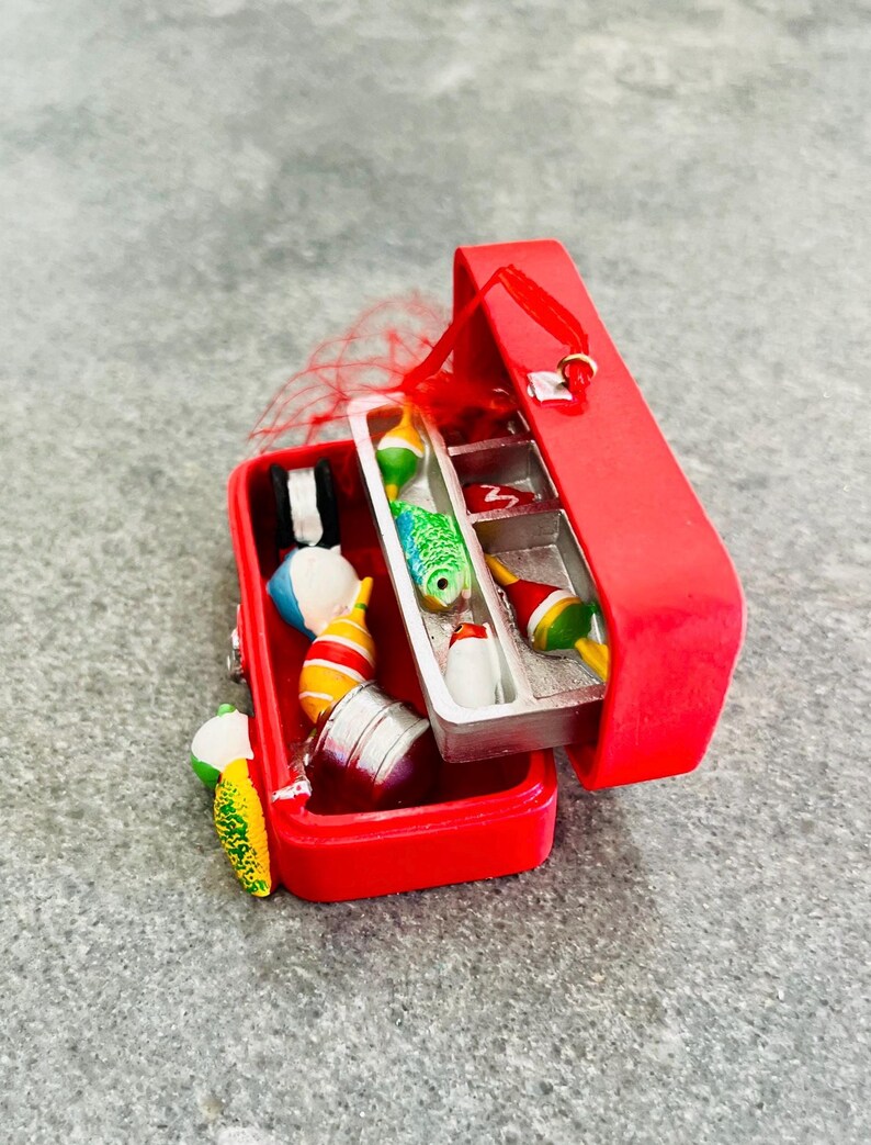 May include: A red miniature tackle box ornament, open to reveal colorful fishing bobbers and lures. The box is rectangular with a hinged lid and a small red ribbon for hanging. The lures include a green and blue fish, a white and red bobber, and a yellow and green lure.